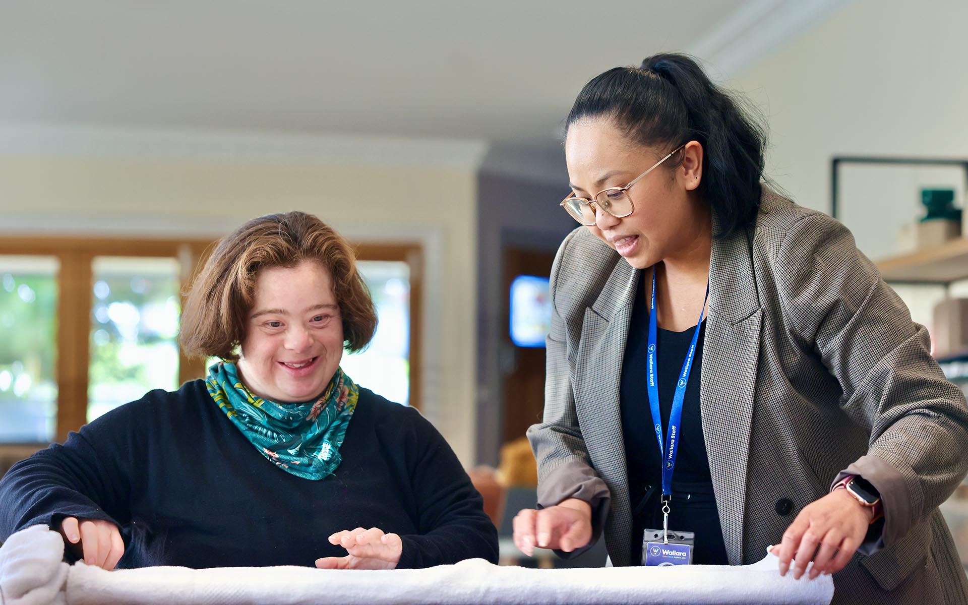 A Wallara staff member helping a participant fold towels in a home environment.