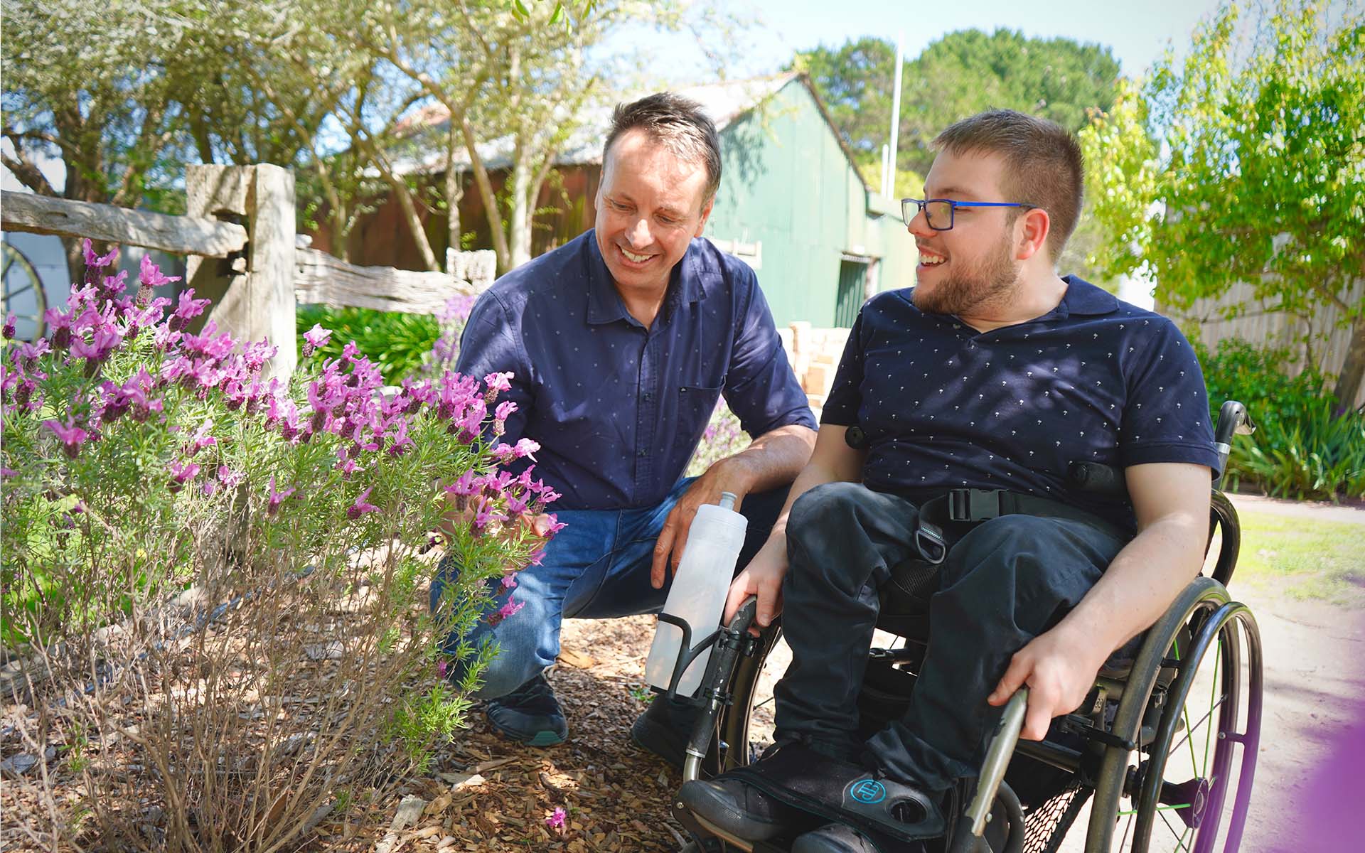 A Wallara participant using a wheelchair and a support worker spend time together in a garden, looking at flowering plants.